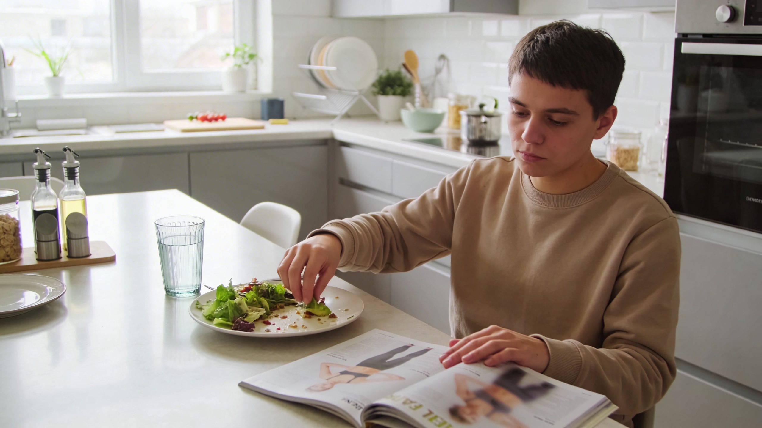 A person sitting at a kitchen island eating a healthy salad while reading a fitness magazine.