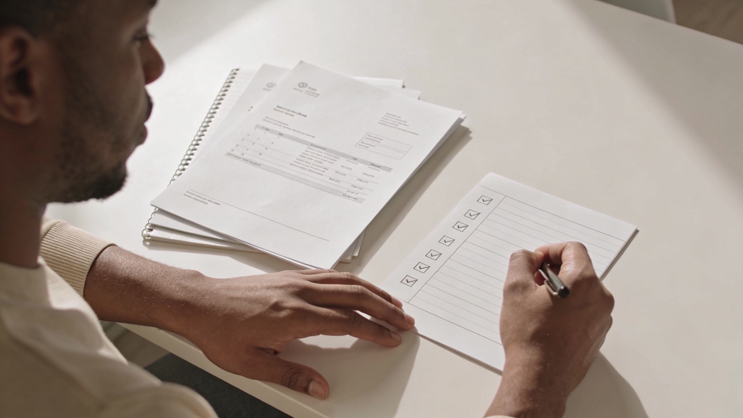 A person writing on a checklist paper while reviewing documents on a desk with a pen.